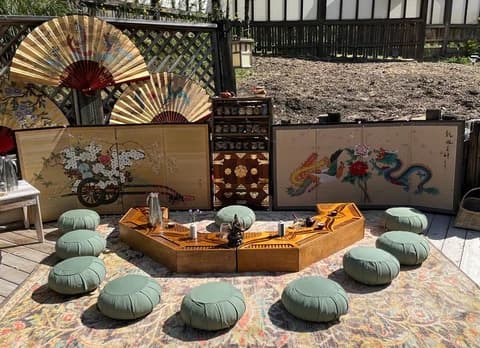 A serene tea lounge with a wooden tea table, sage cushions, and ornate fans